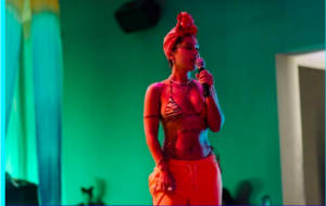 A black woman bathed in red light holding a microphone , the background behind them is green , they are wearing a headscarf, triangle bikini top and trousers