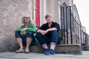 Jeni & kirsty sit on a doorstep in frint of a red door and an iron railing, the are both smiling