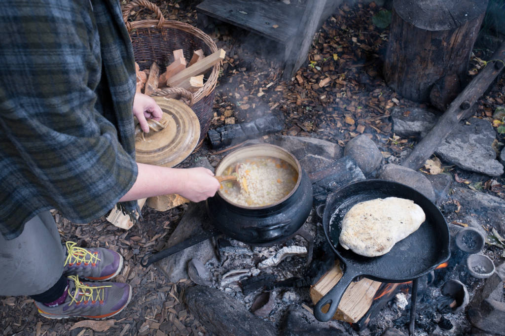 At The Scottish Crannog Centre a person leans over a coal fire to stir some soup in a traditional pot , there is bread to the left and fire kindling to the right