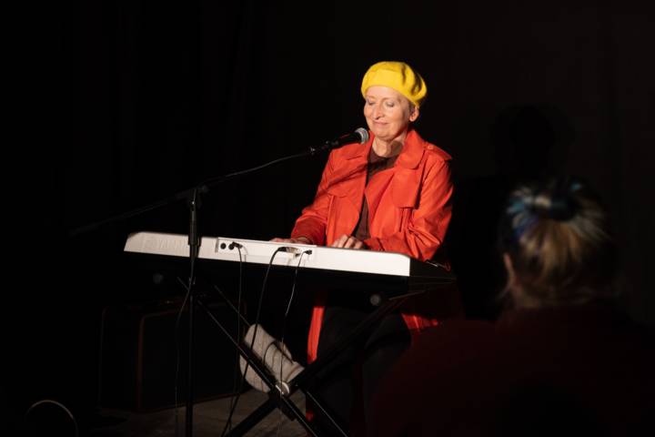 A woman wearing a yellow berret and a red jacket sits at an electronic keyboard in. dark room, performing at Dundee Fringe Festival
