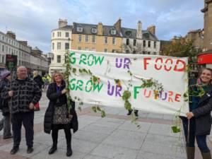two people hold a handmade banner in the city square, it reads, 'grow ur food, grow ur future'