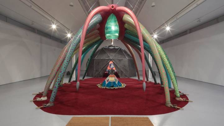 An installation of Rae yen songs exhibition at DCA featuring a round red carpet with a giant inflatable creature with many legs standing over an amorphous sculpture of a person/creature in the middle, the colours are bright and warm