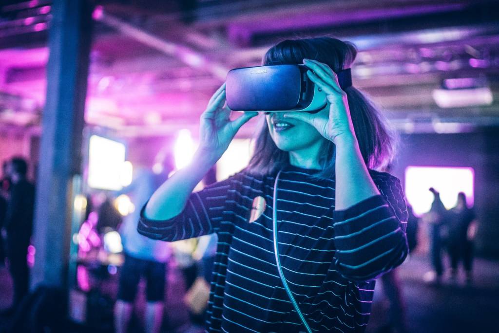 A women with shoulder length hair and a stripey top wears a VR headset - her hands are raised and she holds the headset from each side.