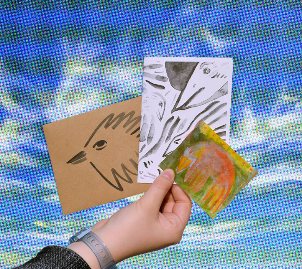 A hand holds three small drawings of birds and animals by artist Maria Tolia against a blue sky with white clouds.