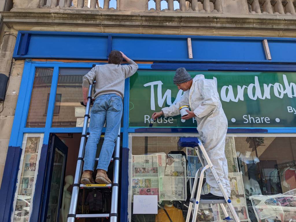 Two people are on ladders outside a shop called The Wardrobe. They are both using dark blue paint to repaint the shop front.