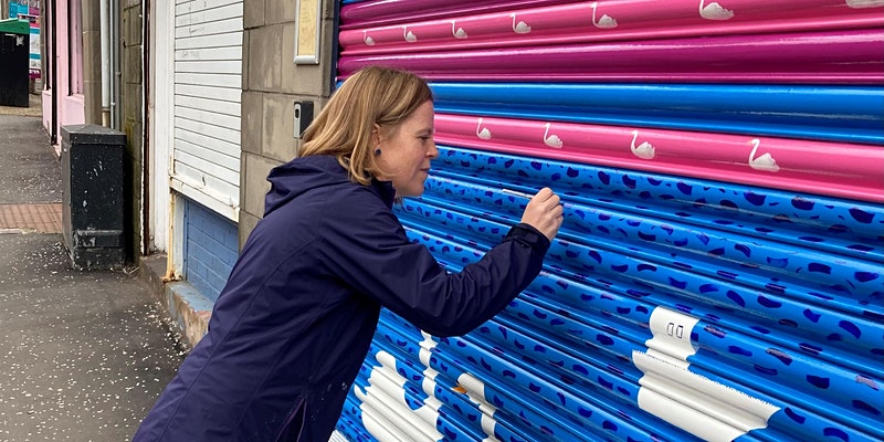 Artist Louise Kirby is painting a shop shutter with patterns and swans on a street in Stobswell.