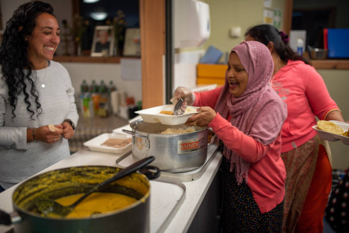 Photograph of a group of three people. A woman in a pink headscarf is serving food from a large cooking pot while two others look on smiling and laughing.