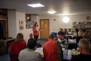 A Photograph. A person stands at the front a room speaking to a large group of people gathered around tables. Some of the people at the tables are eating and drinking.