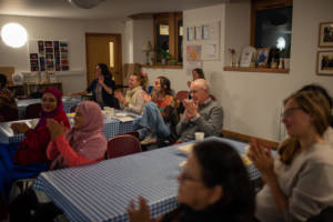 A photograph of people gathered around tables. They are clapping and looking towards the front of the room.