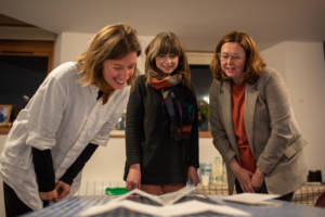 A photograph of 3 people looking at some drawings laid out on a table.