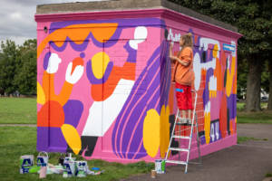 A photograph of artist Zofia painting her colourful mural on the Fair Growing Green maintenance building.