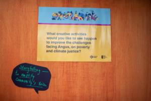Photograph of two signs on a wooden door. The top sign is blue, pink and yellow and shows the CULTIVATE ident above black text which reads: 'What creative activities would you like to see happen to improve the challenges facing Angus, on poverty and climate justice?' The sign below is black and shaped like a speech bubble, green handwritten text on it reads: 'Storytelling - to amplify community voices'