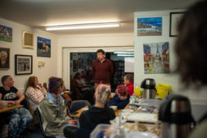 Photograph of a man in a dark red jumper speaking to a group of people gathered around tables. A variety of paintings are hung on the white walls and a buffet of food is laid out in the foreground.