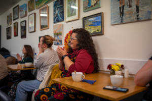 A photograph of a group of people sitting at small coffee tables and clapping. Behind them a variety of paintings and photographs hang on the wall.
