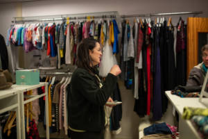 A woman with dark brown hair and wearing a black fleece is stood in a clothing shop. Clothes are hung on rails behind her and displayed on white ables around her.