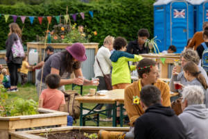 Photograph of a busy community garden. Groups of people and families are gathered around picnic tables in the garden.