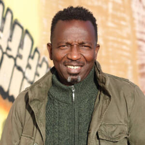 Amadu smiles at the camera while stood in front of a washed out orange and yellow painted wall. He has dark cropped, curly hair and a goatee. He is wearing a khaki jacket over a zip-up dark green jumper.