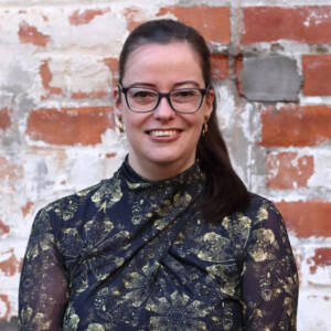 Angie smiles at the camera while stood in front of a red brick wall. She has her dark hair tied back into a ponytail and has dark rimmed glasses. She is wearing a black high neck top with a gold floral pattern.