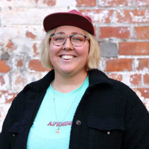 Jaz smiles at the camera while stood in front of a worn brick wall. She has short blonde hair and glasses. She is wearing a burgundy baseball cap, a black jacket and a bright turquoise tshirt.