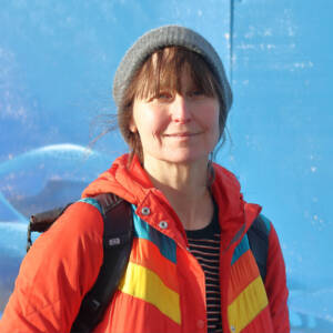 Lu smiles at the camera while stood in front of a bright blue painted wall. She is wearing a grey beanie hat with her fringe poking out, and a bright orange puffer jacket with a blue and yellow stripe across the chest.