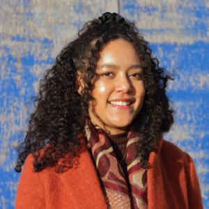 Shona smiles at the camera while stood in front of a blue painted wall. She has shoulder length dark curly hair and is wearing a burnt orange coat and red/brown patterned scarf.