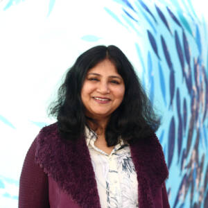 Vinishree smiles at the camera while standing in front of a mural of a school of fish. She has shoulder length hair and is a wearing a burgundy jacket with a fluffy collar.
