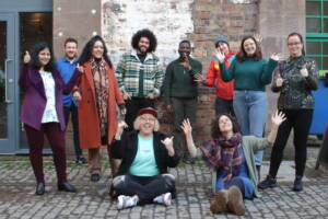 The six Creative Practitioners stand against a brick wall in a cobbled street. They are joined by three members of the Creative Dundee Team and Matt Hickman from Culture Collective. They are all smiling and waving at the camera.