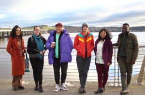 The six creative practitioners, five women and one man, stand in a row on the Dundee waterfront path with the Tay and Tay road bridge behind them.