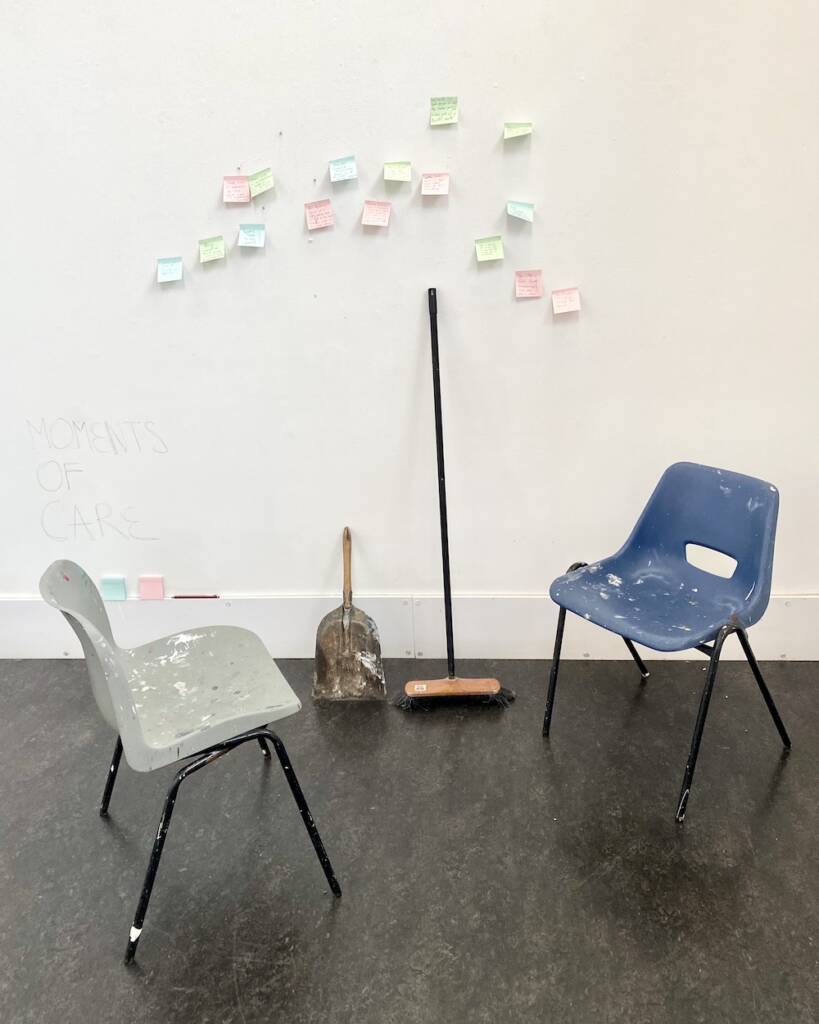Two paint-stained plastic chairs face one another. Leaning on the wall next to them are a dustpan and brush. On the wall above these are 16 colourful sticky-notes.