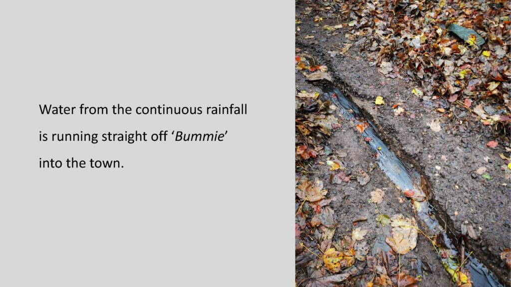 Photograph of water flowing through a widening crack in the ground. Black text to the left of the image reads: Water from the continuous rainfall is running straight off 'Bummie' into the town.