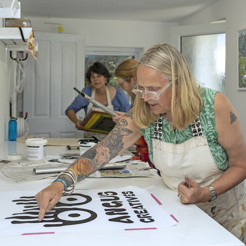 Three people wearing aprons stand at a long-bench doing screen-printing. The person in the foreground, with shoulder-length blonde hair, glasses and a sleeve of tattoos, works on a piece that features the Angus Creatives logo.