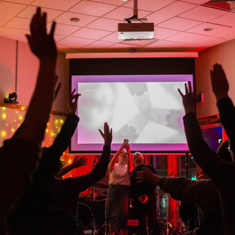 In a darkened room lit by fairy lights and red lighting, two people stand in front of a projected screen taking a phone photo of the crowd who have their hands raised in the air.