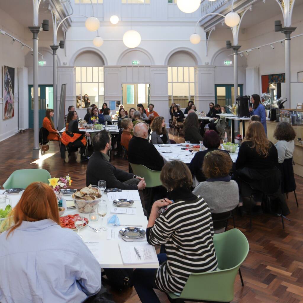 In a wide-open hall in a building, groups of people sit around tables, looking in the direction of a person addressing the room. The tables have plates of food and flowers alongside notebooks and pens.