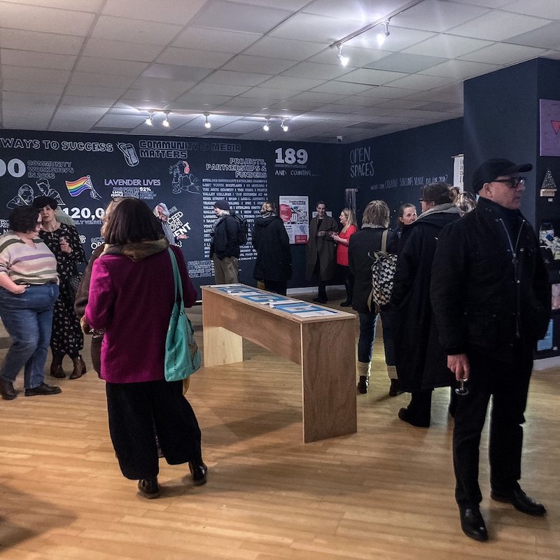 A room full of people at a community exhibition. Dark blue walls are covered in white text and illustrations detailing figures and information about workshops, project funding and participation.