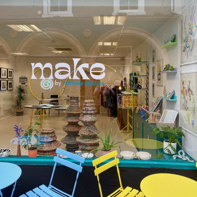 Exterior photo of a shop – the window vinyl reads 'Make by narture studios', above brightly coloured outdoor tables and chairs. Through the window is a bright white space with framed artworks on the walls and prints and sculpture placed around the room.