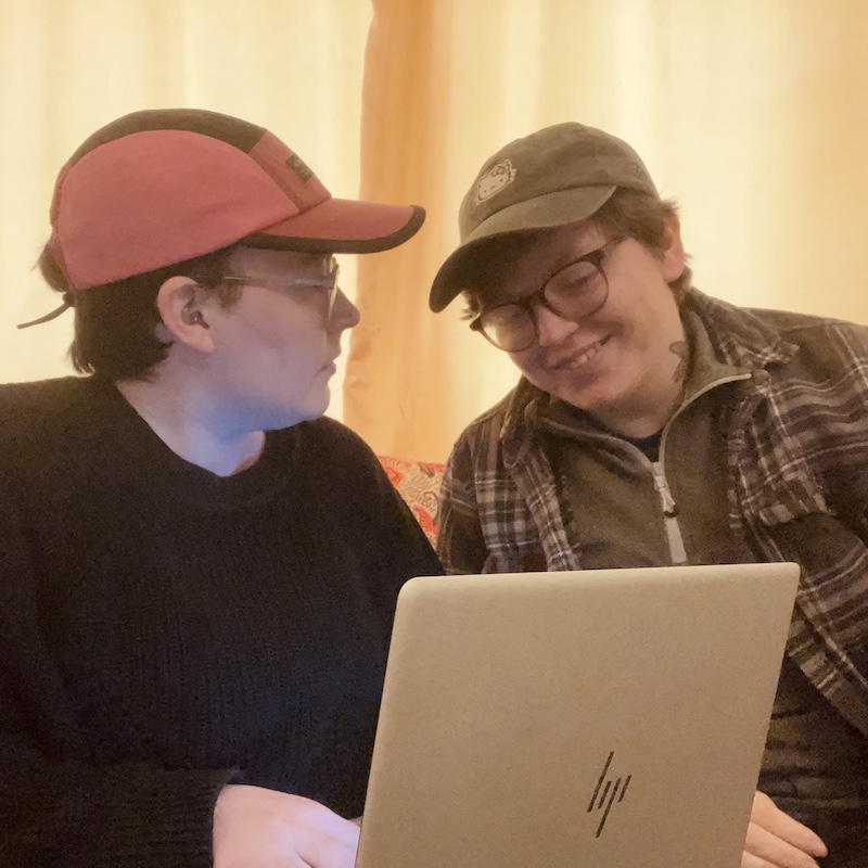 Two people sit together at a laptop. They both wear glasses and baseball caps. One person is looking at the screen and smiling; the other, who holds the laptop, is looking at the other person.