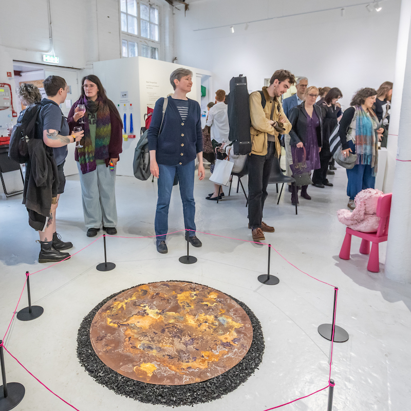 A busy room of people at an exhibition. They're in a large white space, chatting together and looking at artworks that include a circular sculpture on the ground that's cordoned off by a low-red rope.