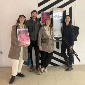Becca and Su of dundee radio club pose with Lizzie and Luke of Volk Gallery, a vending machine gallery mounted on the wall of a small shipping centre, seen behind them.